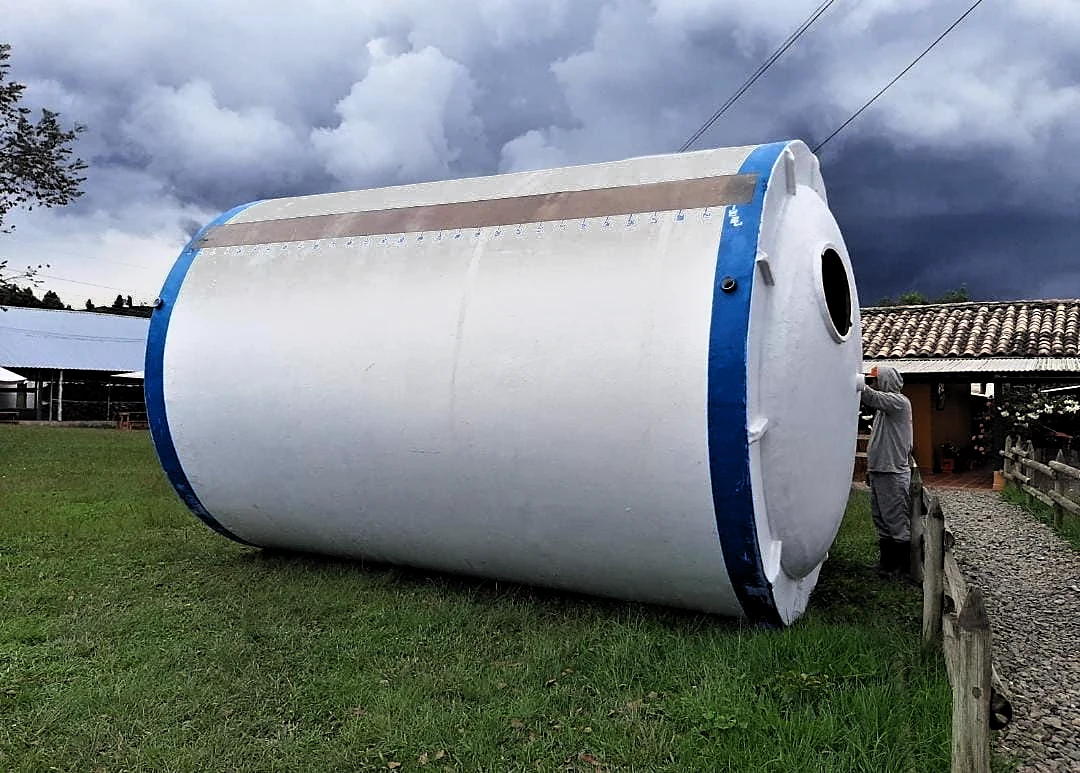 Tanque horizontal en fibra de vidrio terminado, con la escala graduada rotulada sobre el lomo, manhole y orejas de izaje; un operario de pie al lado da la escala del cuerpo.